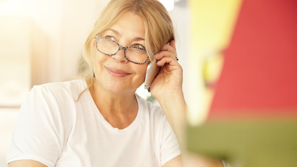 Beautiful blonde Caucasian elderly female wearing glasses having phone conversation with her old friend using electronic device, discussing latest news with cute smile on her face, sitting at home